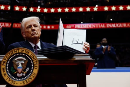 man in suit holds up signed document while seated at desk with presidential seal