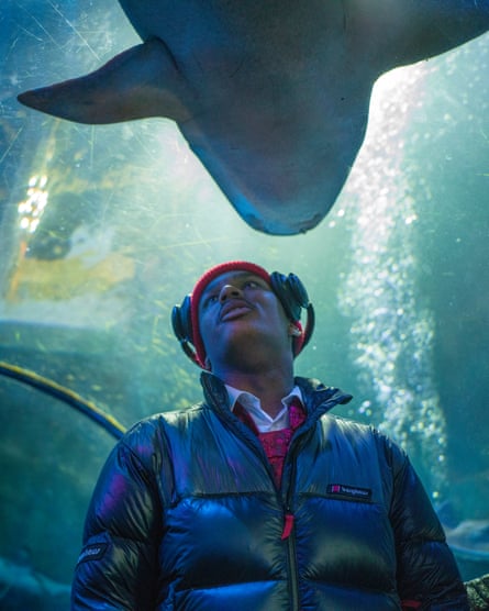 Shot from a low angle, a young African-Caribbean man in a puffer jacket, beanie hat and headphones in an aquarium with a shark edging into view above his head.