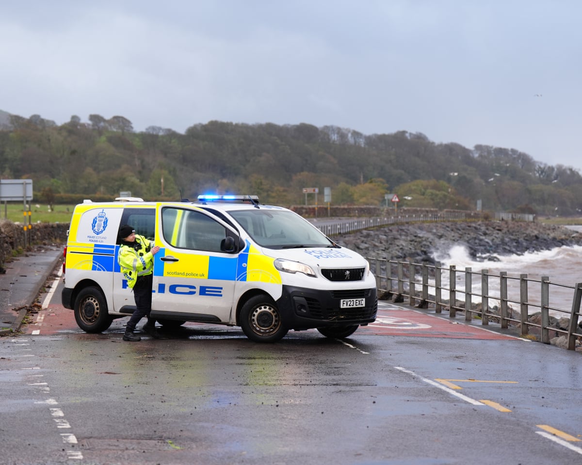 A police officer struggles against the wind to close the door of the police van on a closed coastal road near Skelmorlie