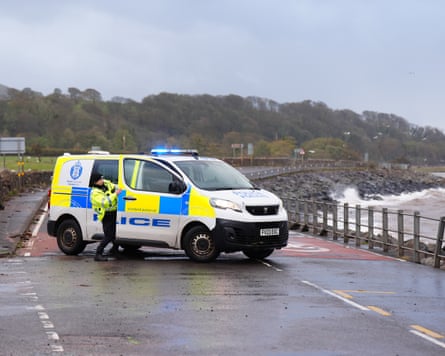 A police officer struggles against the wind to close the door of the police van on a closed coastal road near Skelmorlie