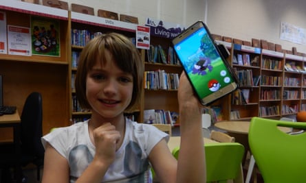 Young girl sitting in library holds up phone displaying Pokemon Go game.