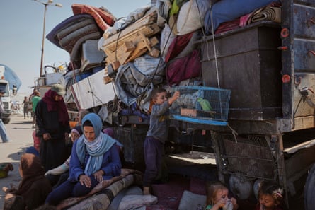 women and children sit and stand beside an overloaded truck which is piled high with household goods, bedding and furniture; a boy is holding a cage containing a green parakeet.