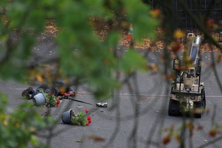 A bomb disposal robot works by a body lying in a street; it is seen through tree branches. There are upturned flower pots around the body.