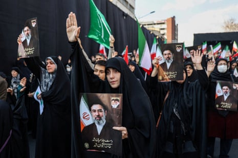 Women in all black and wearing dark hijabs hold up photos of the new supreme leader, Ayatollah Mojtaba Khamenei. Some women are also holding a hand up to the sky, as if in praise.