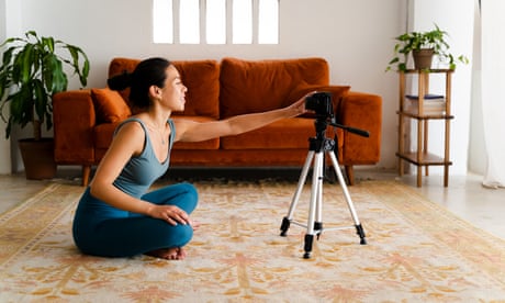 Yoga instructor in blue sportswear preparing to film a yoga video tutorial with a camera on tripod while sitting on carpet in her living room at home.