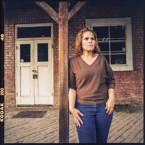 A woman stands in front of an old brick building, staring off into the distance.