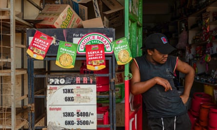 A man in a vest leans against a stack of shop crates hung with a banner advertising instant noodles