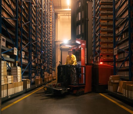 A man in a small forklift truck drives between two very high racks of shelving containing archive boxes in a warehouse
