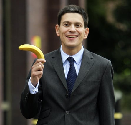 David Miliband wearing a grey suit and a blue shirt and tie, and holding an unpeeled banana
