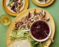 A platter displays pancakes, shredded chicken, cucumber strips, and a bowl of dark plum sauce alongside assembled wraps
