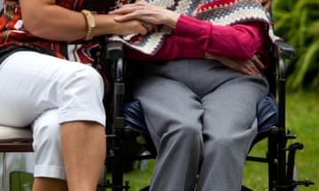 A female carer holds the hand of an elderly woman in a wheelchair