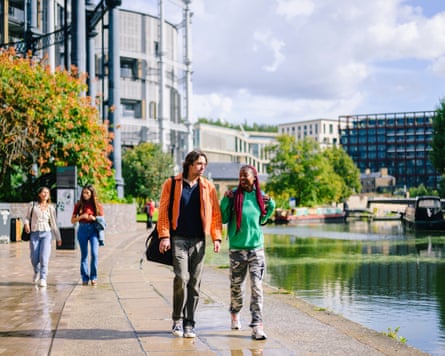 Man and woman walking on path alongside a canal, talking, with city buildings in background
