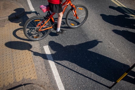 A girl on a bike crossing a road. The shadow of a lollipop person is in the foreground of the picture