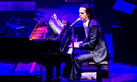 Nick Cave sits at a grand piano, singing into a microphone, at the Hammersmith Apollo in London. 