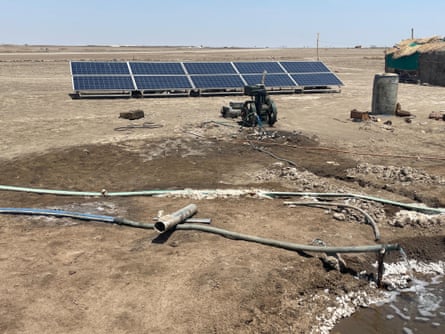A line of solar panels behind a small generator behind a depression in the ground into which a hose is pouring water.