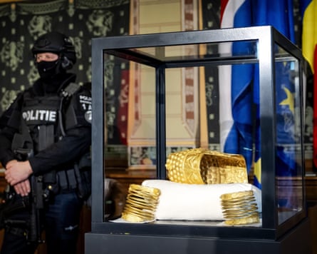 A police officer wearing a balaclava and holding a gun stands beside a display case containing the helmet