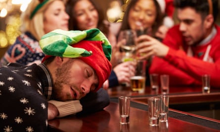 young man wearing a green and red elf hat and Christmas jumper rests his head with his eyes closed on a bar next to empty shot glasses while a group of people in the background are laughing and drinking