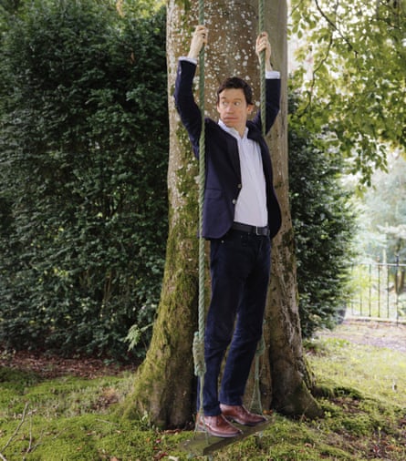 Rory Stewart wearing a navy suit and white shirt and standing on a swing hanging from a tree in a garden