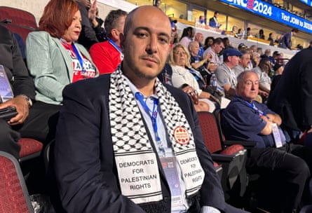 Man seated in arena looking at camera, wearing keffiyeh that says Democrats for Palestinian Rights.