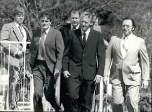 A thin older white man among a group of white officials walking through a garden
