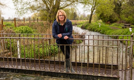Katherine Foxhall in outdoor clothes and wellies standing on a small bridge over a stream with a water depth gauge to her right