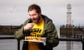 Daniel Gray putting a chip in his mouth with one hand and holding a packet of fish and chips on the other, wearing a yellow T-shirt saying 'fish and chips' and a khaki trenchcoat, with the sea, a lighthouse and a grey sky behind him