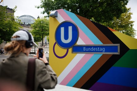 Sign at the entrance to Bundestag station, painted in the Pride flag colours; a person is in front of it taking a photo and a domed parliament building is in the background.