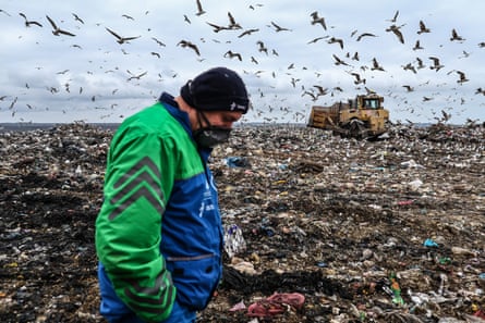 Man walks on a landfill site, while a bulldozer shifts waste in the background.