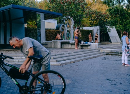 People stand around a paved area in the park. A man attaches something to his bike and people in the background are drying off after being in the water