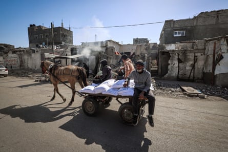 Three people sit on a horse-drawn cart loaded with several sacks of flour in an area of partly destroyed buildings