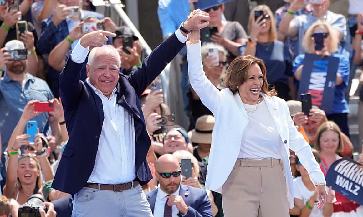 Kamala Harris and Tim Walz smiling and holding hands aloft with crowds in background