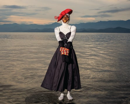 A woman wrapped in bandages, wearing a dress and a red hat, is bound by brightly coloured material. She appears to stand on water in a lake with hills in the background
