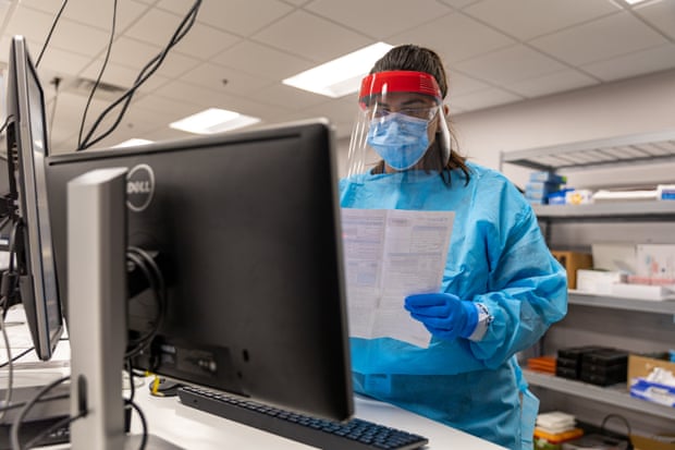 person wearing mask and face shield holds document in front of computer monitor