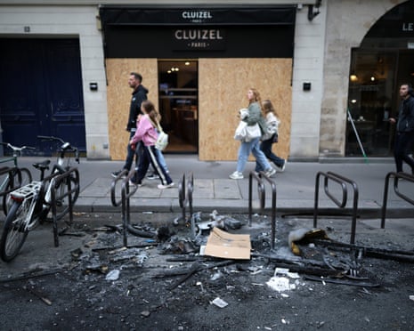 Pedestrians walk past a Paris shop with boarded-up windows, beside burnt protest debris