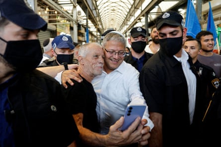 Itamar Ben-Gvir embraces a supporter for a selfie, surrounded by security officers at a market