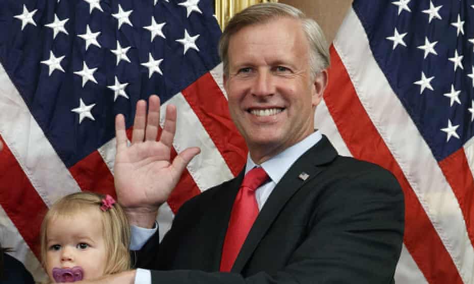 A man in a black suit with a red tie waves to the camera against a backdrop of American flags.