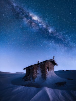 A brick alpine hut is partially covered in snow on the top of a mountain. The night sky above the hut is dotted with millions of stars and what look like clouds making up the Milky Way
