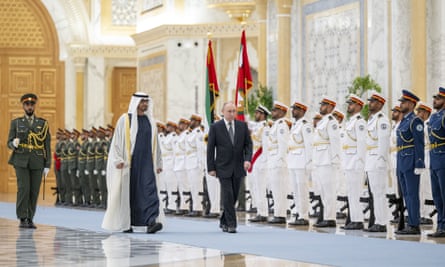 Mohamed bin Zayed Al Nahyan welcomes Vladimir Putin with an official ceremony at Qasr Al Watan in Abu Dhabi – they are both walking on a blue carpet in a grand reception room in front of a line of men in military uniforms, standing to attention