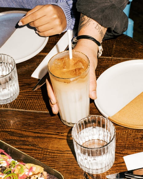 A person holds an iced coffee drink with a straw at a restaurant table with plates and food