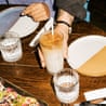 A person holds an iced coffee drink with a straw at a restaurant table with plates and food
