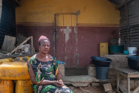 A sad-looking woman sitting outside a red and yellow building, around which are piled up yellow water butts and buckets.
