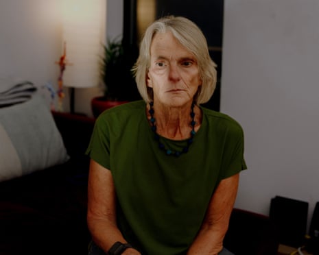 A moody, dark photo of an older white woman with chin-length blond hair, wearing a dressy dark green T-shirt and black necklace.