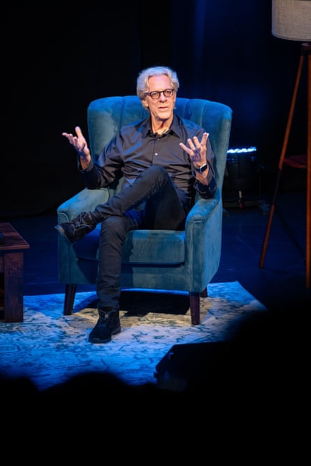 A man in dark pants and a dark shirt wearing dark-rimmed glasses sitting in a blue velvet armchair on stage, gesturing with his hands.