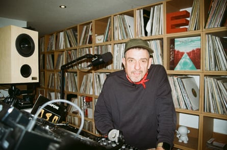 A man wearing a black sweatshirt, red neckerchief and green cap stands nect to DJ decks with a wall of vinyl behind