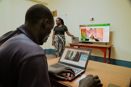A man works on a computer while a woman leads a lesson at the front of the room