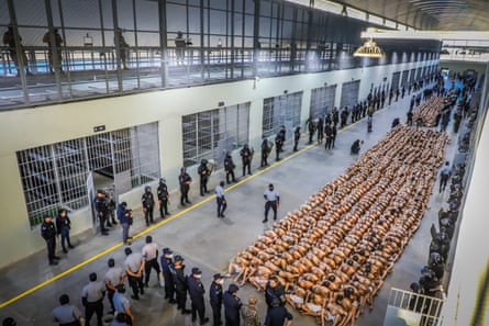 Aerial shot of a indoor prison area, where hundreds of inmates sit packed in rows closely together, wearing only underwear, heads bowed, as standing prison officers look on.