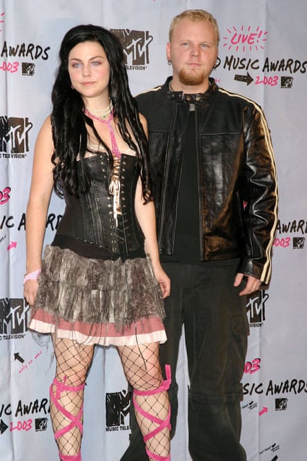 A woman with long black hair, wearing a black waistcoat, a skirt, fishnet tights and pink shoe laces up to her knees. She is standing beside a man with short blond hair wearing a leather jacket.