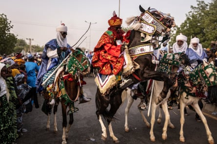 Members of the royal guard wear ceremonial robes and turbans as they ride down a street; the horses have decorative saddlecloths and headdresses, and one horse is rearing on its hind legs in front of the camera