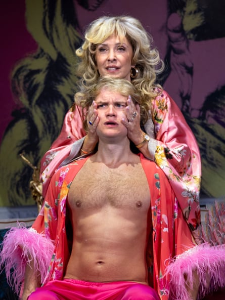 A blonde woman stands above a blonde man who is sitting below her. She is holding his face and they are both dressed in pink robes.