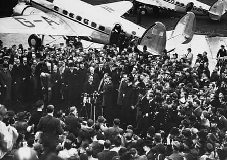 Black and white image of Neville Chamberlain surrounded by a crowd in an airfield. There are planes in the background
